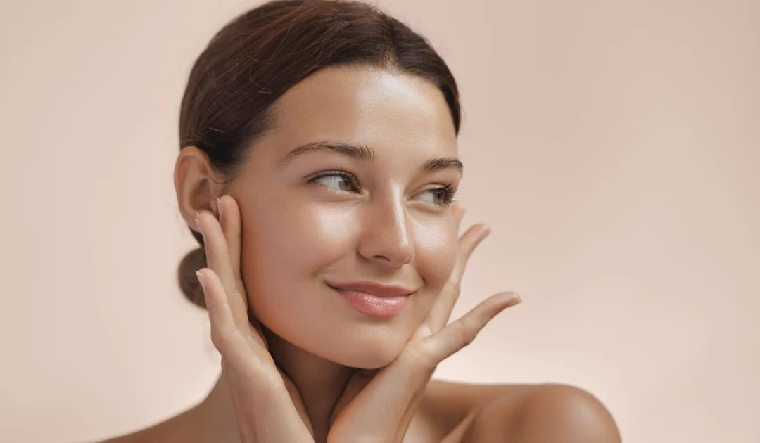 Studio portrait of a woman with glowing skin, hair pulled back, gently touching her face, showcasing Sofwave in Baldwinsville, NY, against a soft light peach background.